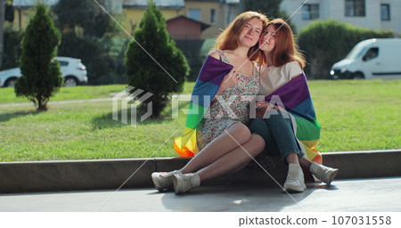 Portrait of two beautiful young women hugging in park under a rainbow flag. Romance and portrait of lesbian couple enjoying. Lifestyle and relationships. LGBT concept. Portrait of two beautiful young women hugging in park under a rainbow flag. Romance and portrait of lesbian couple enjoying. Lifestyle and relationships. LGBT concept. 107031558
