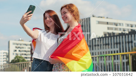 Lesbian couple taking selfie in the city. Two happy girlfriends pose with rainbow flag. Lgbt woman, showing love to each other. Happy lesbian relationships. Lesbian couple taking selfie in the city. Two happy girlfriends pose with rainbow flag. Lgbt woman, showing love to each other. Happy lesbian relationships. 107031642