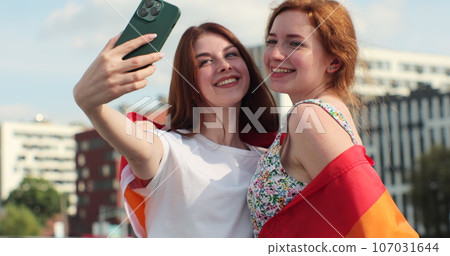 Young lesbian couple taking selfie in the city. Two happy girlfriends pose with rainbow flag. Lgbt woman, showing love to each other. Happy lesbian relationships. Young lesbian couple taking selfie in the city. Two happy girlfriends pose with rainbow flag. Lgbt woman, showing love to each other. Happy lesbian relationships. 107031644