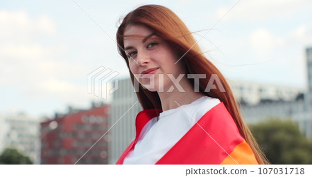 Beautiful young woman with long red hair covering in a rainbow flag. Alone. One. Keeping fist up, covering LGBT flag. Beautiful young woman with long red hair covering in a rainbow flag. Alone. One. Keeping fist up, covering LGBT flag. 107031718