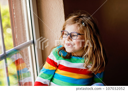 Adorable little preschool girl with eyeglasses sitting by the window. Thoughtful child looking out. Lonely kid. 107032052