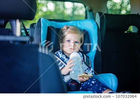 Adorable baby girl with blue eyes sitting in car safety seat. Toddler child going on family vacations and jorney. Smiling happy child during traffic jam, drinking milk from bottle and eating bisquit Adorable baby girl with blue eyes sitting in car safety seat. Toddler child going on family vacations and jorney. Smiling happy child during traffic jam, drinking milk from bottle and eating bisquit 107032064