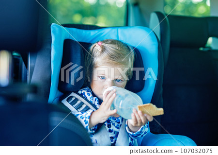 Adorable baby girl with blue eyes sitting in car safety seat. Toddler child going on family vacations and jorney. Smiling happy child during traffic jam, drinking milk from bottle and eating bisquit 107032075