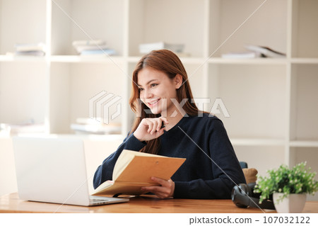 A beautiful caucasian female student is studying remotely. She is sitting at a table at home with a laptop and a notepad and concentrated is watching a video conference, Thinking. A beautiful caucasian female student is studying remotely. She is sitting at a table at home with a laptop and a notepad and concentrated is watching a video conference, Thinking. 107032122
