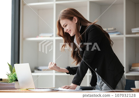 Portrait of Beautiful happy young business woman celebrating success with arms up in front of laptop. Freelancer finished project. 107032128