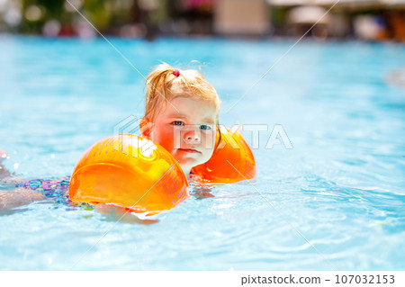 Little toddler girl with protective swimmies playing in outdoor swimming pool by sunset. Baby Child learning to swim in outdoor pool, splashing with water, laughing and having fun. Family vacations. 107032153