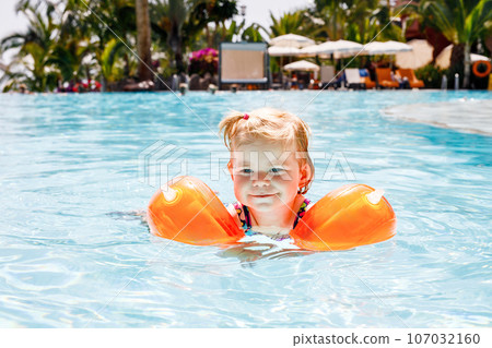 Little toddler girl with protective swimmies playing in outdoor swimming pool by sunset. Baby Child learning to swim in outdoor pool, splashing with water, laughing and having fun. Family vacations. Little toddler girl with protective swimmies playing in outdoor swimming pool by sunset. Baby Child learning to swim in outdoor pool, splashing with water, laughing and having fun. Family vacations. 107032160