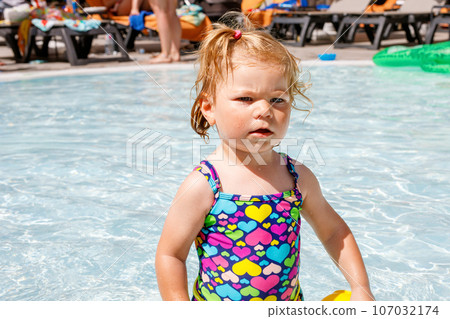 Cute happy little toddler girl in the pool and having fun on family vacations in a hotel resort. Healthy child playing in water. Baby in colorful fashion swimsuit. 107032174