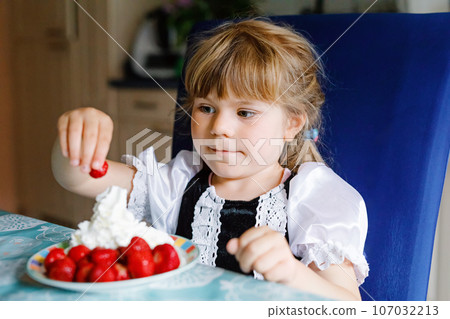 Little preschool toddler girl eating fresh strawberries with whipped cream. Adorable baby child tasting and biting ripe strawberry. Healthy food, childhood and development. 107032213
