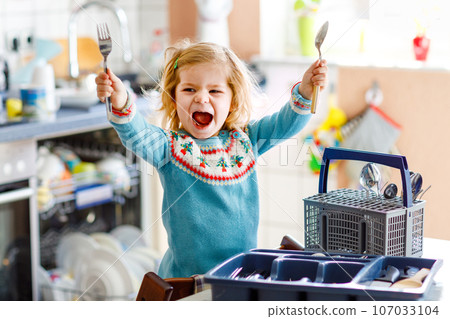 Cute little toddler girl helping in the kitchen with dish washing machine. Happy healthy blonde child sorting knives, forks, spoons, cutlery. Baby having fun with helping housework mother and father. 107033104