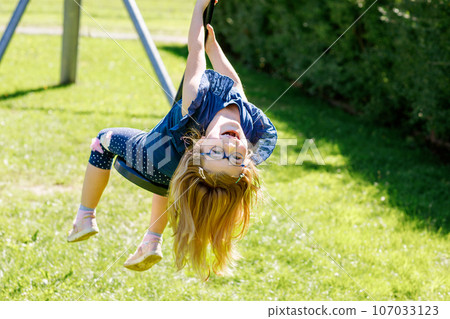 Happy kid girl rids on zip line swing outdoor game play equipment on playground. Child having fun outdoors. Preschool child swinging on summer day. Activity with children. 107033123