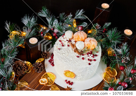 Christmas cake made of chocolate base with cream, decorated with spices, tangerines, pomegranate seeds against the backdrop of Christmas decor 107033698