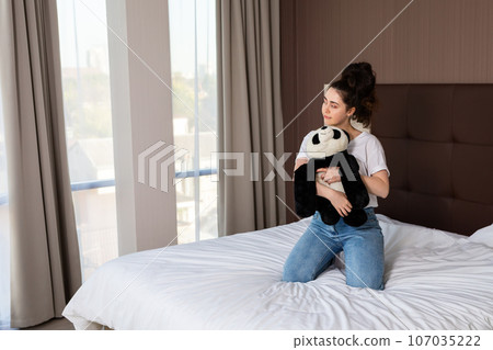 A young woman hugs a stuffed Panda toy in her hands while sitting on the bed and looking out the window. Indoors 107035222