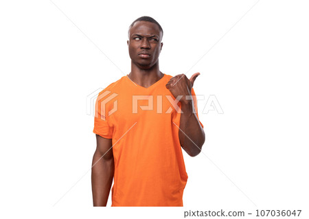 young suspicious african man in orange t-shirt pointing with fingers to the side at advertising 107036047