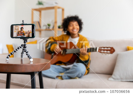 Blogger guitarist. African american girl blogger playing guitar talking to webcam recording vlog. Social media influencer woman streaming at home indoors. Music content creator broadcast tutorial Blogger guitarist. African american girl blogger playing guitar talking to webcam recording vlog. Social media influencer woman streaming at home indoors. Music content creator broadcast tutorial 107036656