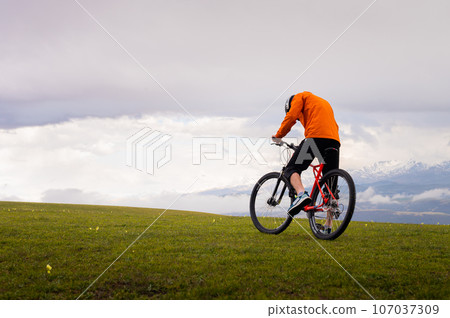 Professional mountain biker on the background of mountains and green hills. Tired mtb rider in full face helmet outdoors Professional mountain biker on the background of mountains and green hills. Tired mtb rider in full face helmet outdoors 107037309
