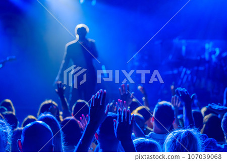 Back view of crowd of unrecognizable people dancing with raised arms while standing near stage during concert of male singer and musician in club 107039068