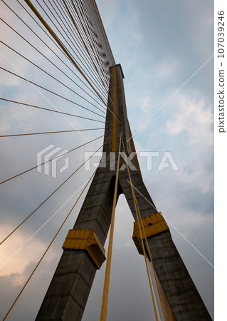 A towering suspension bridge, with concrete pillars and steel cables, cloudy sky. A towering suspension bridge, with concrete pillars and steel cables, cloudy sky. 107039246