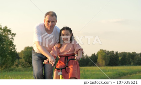 Happy father supports bicycle while little girl learning to ride in green field Happy father supports bicycle while little girl learning to ride in green field 107039621