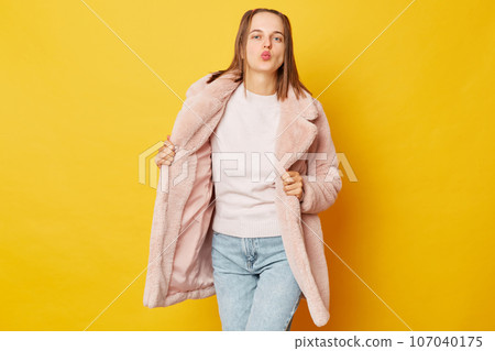 Romantic beautiful brown haired girl wearing pink fur coat with ponytails isolated over yellow background keeps lips folded blows air kiss at camera flirting with boyfriend. Romantic beautiful brown haired girl wearing pink fur coat with ponytails isolated over yellow background keeps lips folded blows air kiss at camera flirting with boyfriend. 107040175