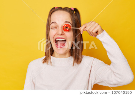 Funny joyful woman with ponytail in jumper holding lollipop isolated over yellow background covering eyes with sugary candy laughing expressing happiness. Funny joyful woman with ponytail in jumper holding lollipop isolated over yellow background covering eyes with sugary candy laughing expressing happiness. 107040190