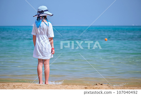 Back view of child girl in white dress and big hat with blue ribbons in long braids standing barefooted on beach enjoying ocean view 107040492