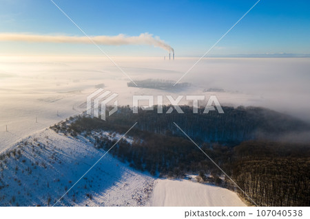 Aerial view of winter landscape with foggy countryside and distant factory pipes emmiting black dirty smoke polluting environment. Aerial view of winter landscape with foggy countryside and distant factory pipes emmiting black dirty smoke polluting environment. 107040538