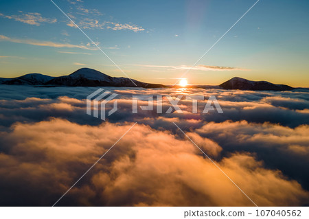 Aerial view of vibrant sunrise over white dense fog with distant dark Carpathian mountains on horizon. 107040562