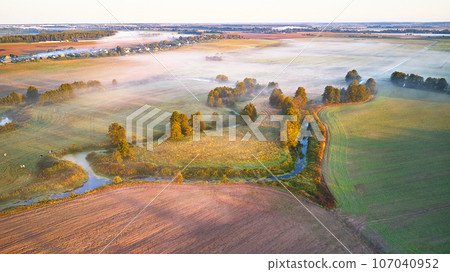 Warm sunny September morning. Autumn beautiful fog landscape. Small river curves in trees. Bright sunny misty riverside. Scenic view of rural sunrise. Village in Agriculture fields Warm sunny September morning. Autumn beautiful fog landscape. Small river curves in trees. Bright sunny misty riverside. Scenic view of rural sunrise. Village in Agriculture fields 107040952