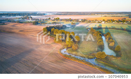 Warm sunny September morning. Autumn beautiful fog landscape. Small river curves in trees. Bright sunny misty riverside. Scenic view of rural sunrise. Village in Agriculture fields 107040953