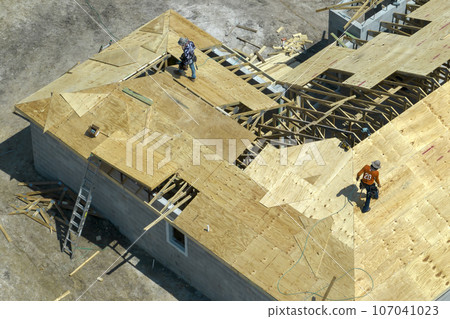 Aerial view of builders working on unfinished residential house with wooden roof frame structure under construction in Florida suburban area Aerial view of builders working on unfinished residential house with wooden roof frame structure under construction in Florida suburban area 107041023