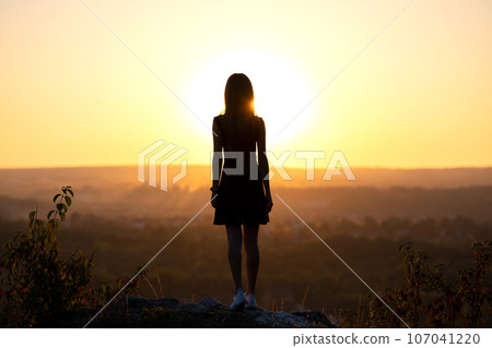 A young woman in summer dress standing outdoors enjoying view of bright yellow sunset. 107041220