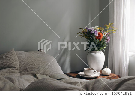 Elegant moody bedroom interior. Cup of coffee, pumpkin on retro wooden bedside table. White ceramic vase. Bouquet of dahlia, cosmos, solidago flowers. Beige muslin cushions, book in bed near window. 107041981