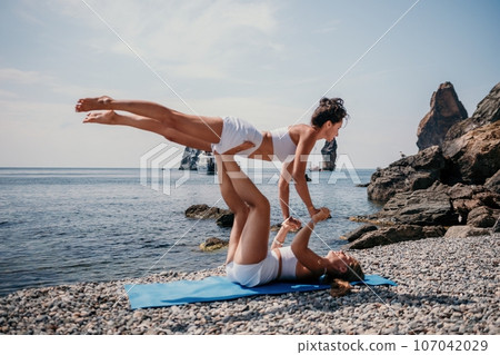 Woman sea yoga. Two Happy women meditating in yoga pose on the beach, ocean and rock mountains. Motivation and inspirational fit and exercising. Healthy lifestyle outdoors in nature, fitness concept. Woman sea yoga. Two Happy women meditating in yoga pose on the beach, ocean and rock mountains. Motivation and inspirational fit and exercising. Healthy lifestyle outdoors in nature, fitness concept. 107042029