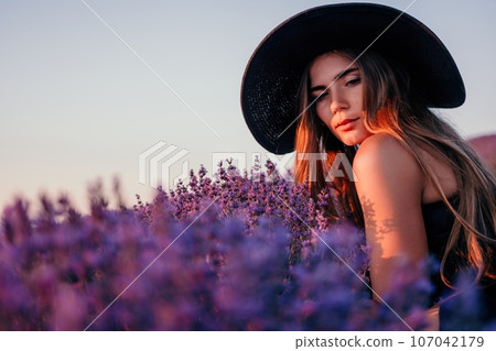 Woman lavender field. Happy carefree woman in black dress and hat with large brim smelling a blooming lavender on sunset. Perfect for inspirational and warm concepts in travel and wanderlust. Close up 107042179
