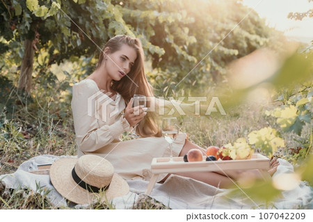 Woman picnic vineyard. Happy woman with a glass of wine at a picnic in the vineyard, wine tasting at sunset and open nature in the summer. Romantic dinner, fruit and wine. 107042209