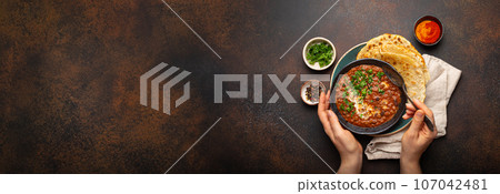 Female hands holding a bowl, eating traditional Indian Punjabi dish Dal makhani with lentils and beans served with naan flat bread, fresh cilantro on brown concrete rustic table top view. Copy space 107042481
