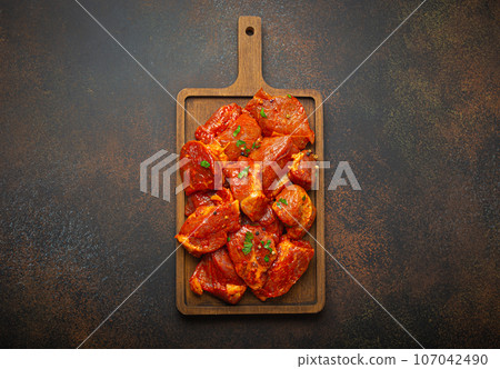 Raw uncooked chopped pieces of pork marinated with seasonings and parsley on wooden cutting board top view on dark rustic background. Cooking meal with marinated pork fillet pieces 107042490