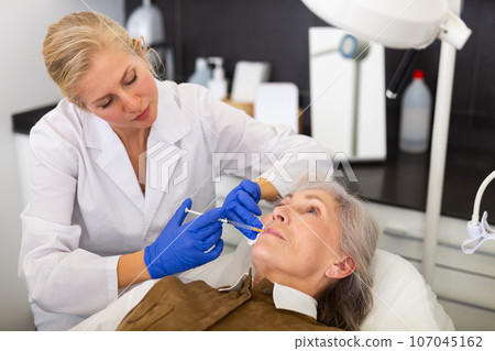 Female cosmetologist injecting filler to correct face contour to senior woman 107045162