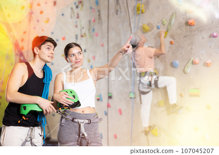 Positive young couple standing with blue safety belts and helmet before practicing rock-climbing on climbing wall in adventure park Positive young couple standing with blue safety belts and helmet before practicing rock-climbing on climbing wall in adventure park 107045207