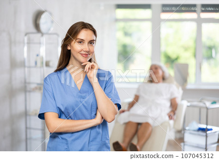 Smiling senior woman doctor in uniform with woman patient lying behind in clinic. 107045312