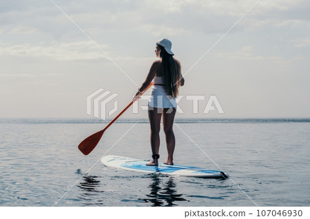 Woman sea sup. Close up portrait of happy young caucasian woman with long hair looking at camera and smiling. Cute woman portrait in bikini posing on sup board in the sea Woman sea sup. Close up portrait of happy young caucasian woman with long hair looking at camera and smiling. Cute woman portrait in bikini posing on sup board in the sea 107046930