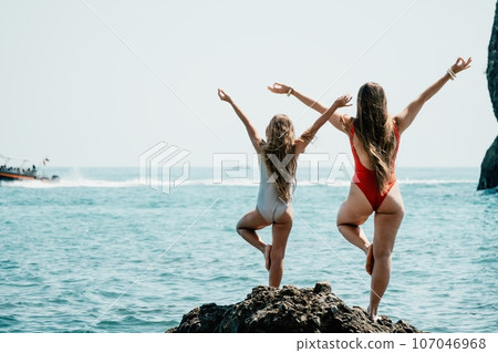 Woman and her daughter practicing balancing yoga pose on one leg up together on rock in the sea. Silhouette mother and daughter doing yoga at beach 107046968