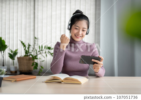 A cheerful Asian woman showing her fist and enjoying watching an online game show on her smartphone. A cheerful Asian woman showing her fist and enjoying watching an online game show on her smartphone. 107051736