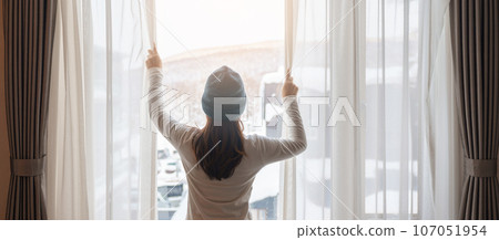 young woman in sweater looking through the window in winter season, happy female rising arms and stretching after waking at apartment or home in the morning. Lifestyle and Relaxing concepts 107051954