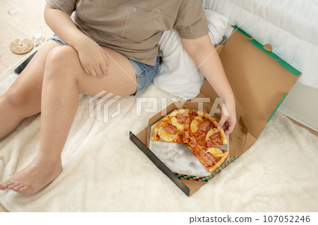 Cropped top view image of a curvy plus-size woman in comfy clothes eating pizza in her bedroom. Cropped top view image of a curvy plus-size woman in comfy clothes eating pizza in her bedroom. 107052246
