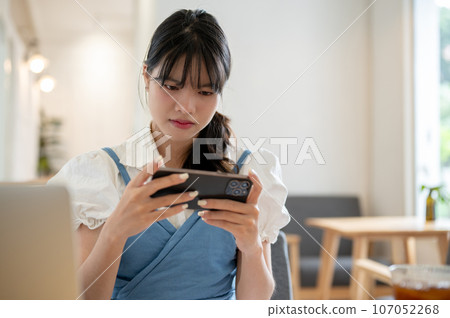A focused Asian woman is playing a mobile game through her phone while sitting in a coffee shop. 107052268