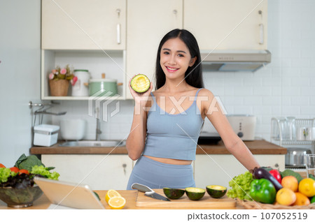 A woman shows an avocado to the camera while she prepares her healthy meals in the kitchen. A woman shows an avocado to the camera while she prepares her healthy meals in the kitchen. 107052519