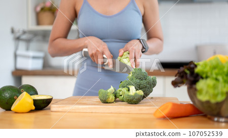 A fit woman in gym clothes is cutting broccoli on a chopping board, preparing her healthy breakfast. A fit woman in gym clothes is cutting broccoli on a chopping board, preparing her healthy breakfast. 107052539