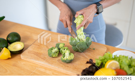 A fit woman in gym clothes is cutting broccoli on a chopping board, preparing her healthy breakfast. A fit woman in gym clothes is cutting broccoli on a chopping board, preparing her healthy breakfast. 107052541
